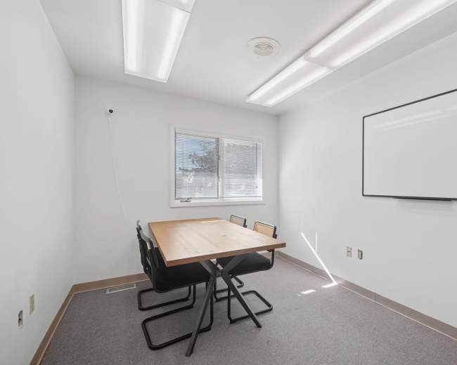 The image shows a small, minimalist meeting room with a rectangular table and four black chairs, a whiteboard on the wall, and a window letting in natural light.