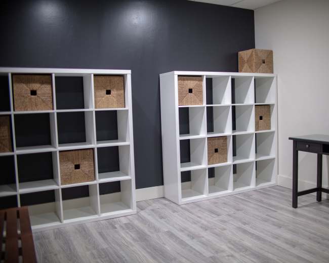 The image shows a simple room with two white shelving units, each containing woven storage boxes, and a black vanity table with a round mirror.