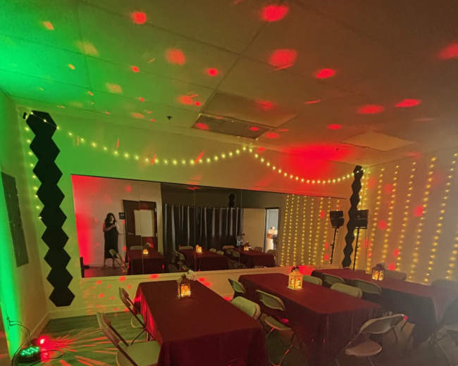 The image shows a dimly lit room set up for an event, featuring several tables covered with maroon tablecloths, decorative lighting along the walls, and a mirrored wall reflecting the space.
