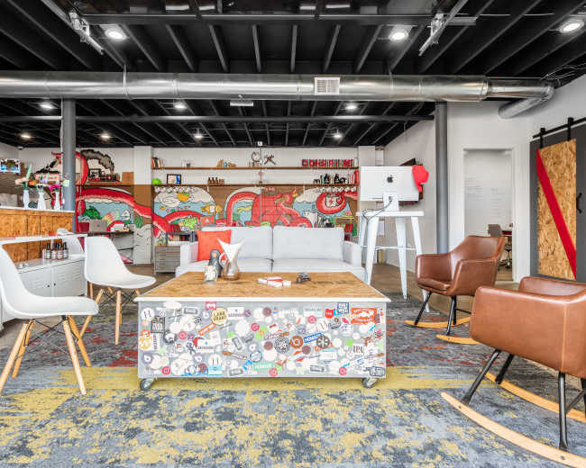 The image shows a modern office space with a gray sofa, wooden coffee table covered in stickers, and brown armchairs, set against colorful wall murals.