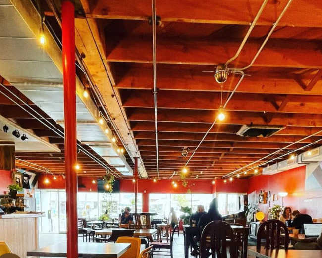 The image shows a brightly lit café with wooden ceilings, colorful wall murals, and patrons sitting at tables while others work on laptops.