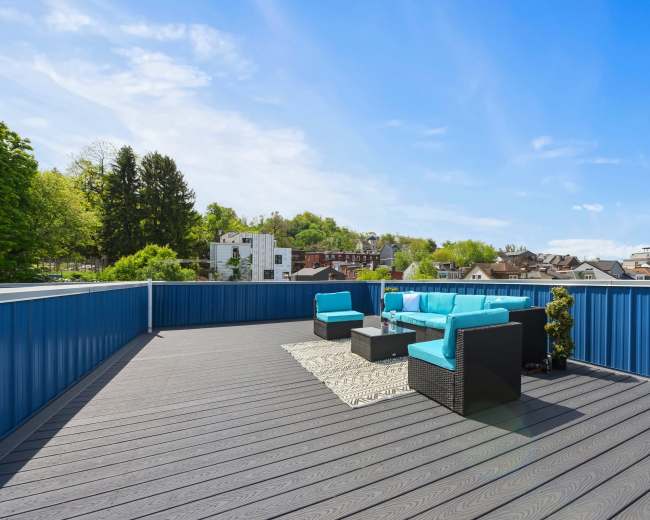 A rooftop terrace features a turquoise seating area surrounded by trees and buildings under a clear blue sky.