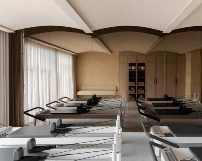 The image shows a modern fitness studio equipped with several reformer machines arranged in rows, with large windows allowing natural light to enter.