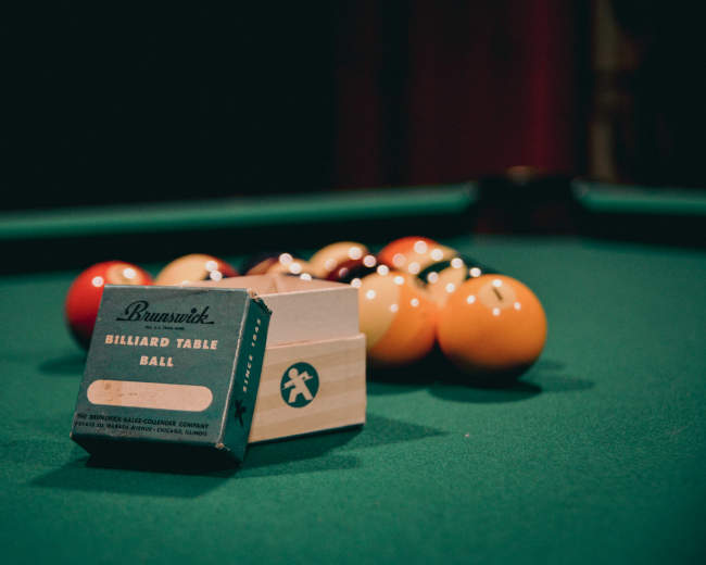 A box of Brunswick billiard table balls sits next to a set of colorful pool balls arranged on a green felt table.