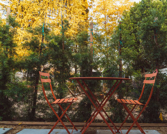 A small round table with two red chairs is set against a backdrop of green shrubs and trees.