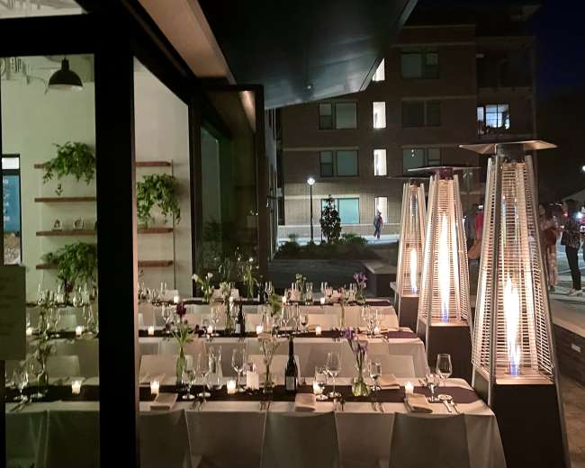 A well-set outdoor dining area of a restaurant at night, featuring tables adorned with white tablecloths, candles, and plants, alongside large outdoor heaters.