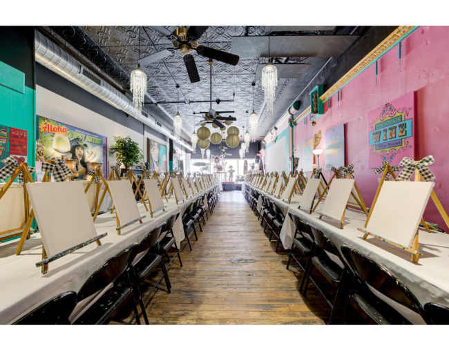 The image shows a brightly lit art studio with a long table set up for painting, featuring empty easels and white tablecloths, lined with chairs on either side.