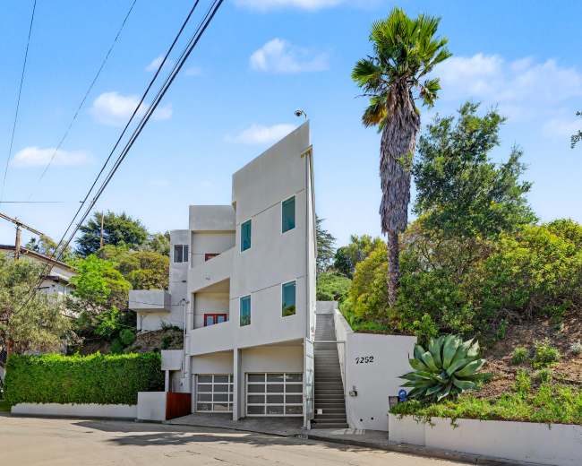 A modern, three-story white building with large windows and a palm tree is situated beside a sloped green landscape and a quiet street.