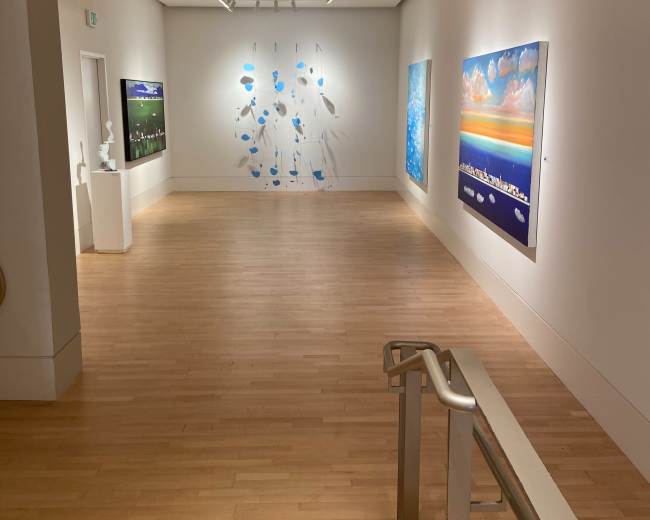 The image shows a spacious art gallery featuring white walls adorned with colorful paintings and suspended art pieces, as well as wooden flooring and a staircase in the foreground.