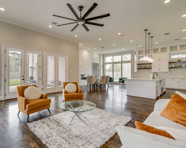 The image shows a spacious living room with a modern open layout featuring a kitchen, dining area, and large windows overlooking a backyard.