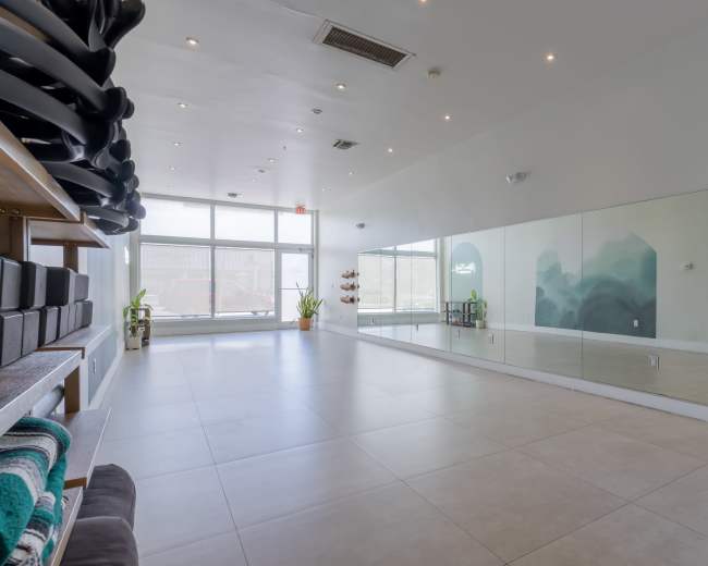 The image shows a spacious, well-lit studio with a large mirror and minimal furnishings, including shelves with yoga accessories.