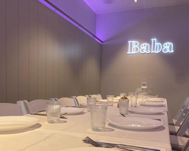A long dining table is set with white plates and glasses, featuring the word "Baba" illuminated on the wall in soft light.