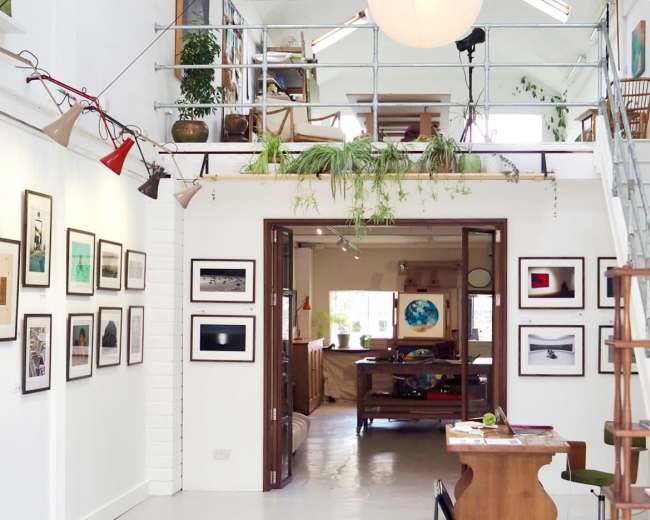 The image shows an art studio with white walls adorned with framed photographs, leading into a spacious area featuring wooden furniture and plants.