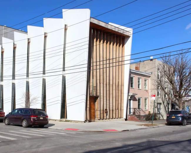 The image shows a modern building with a striking white facade and wooden accents, situated at a street corner next to traditional houses and parked cars.