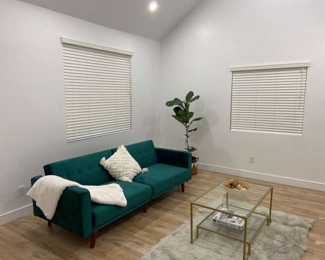 The image shows a modern living room with a teal couch, a coffee table, and two windows covered with white blinds.