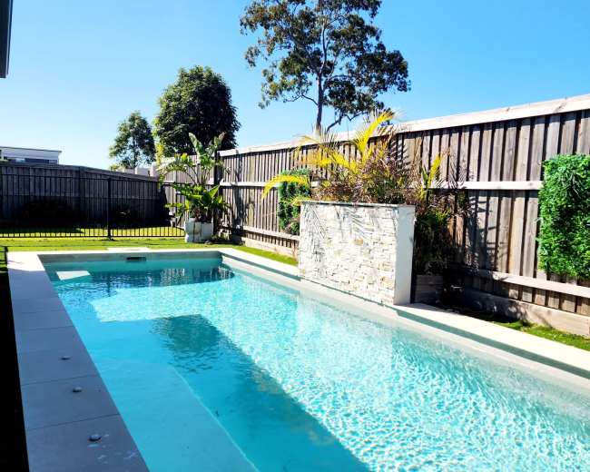 A rectangular swimming pool is surrounded by landscaped greenery and wooden fencing under a clear blue sky.