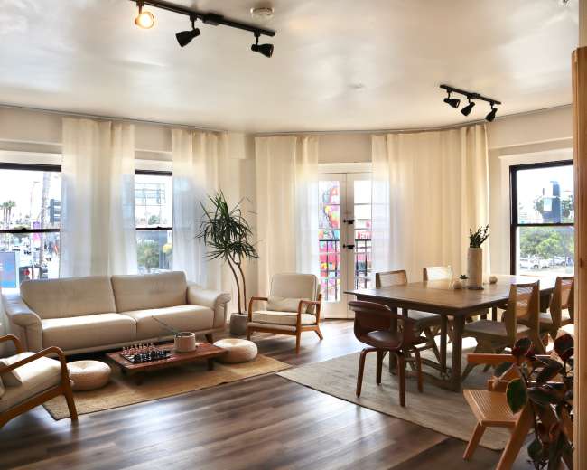 The image shows a bright, modern living room with a white sofa, wooden dining table, and large windows draped with sheer curtains.