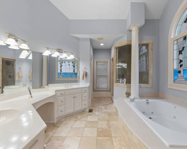 The image shows a spacious bathroom featuring a large whirlpool tub, a double vanity with light fixtures, and a curved shower enclosure with stained glass accents.