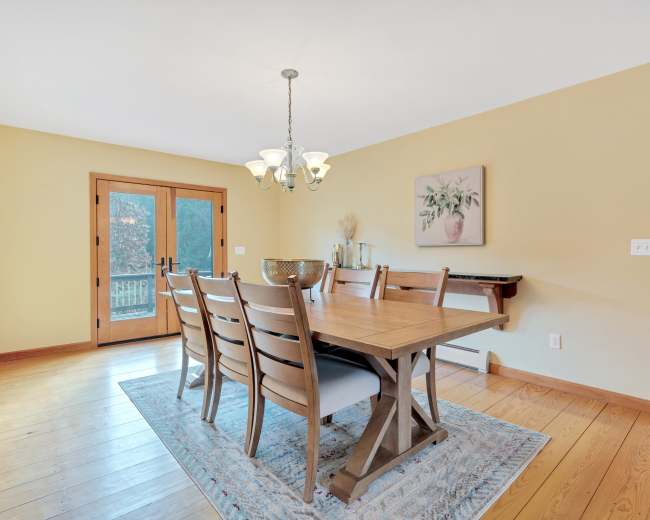 A dining room features a wooden table with six chairs, a chandelier above, and double glass doors leading to an outdoor area.