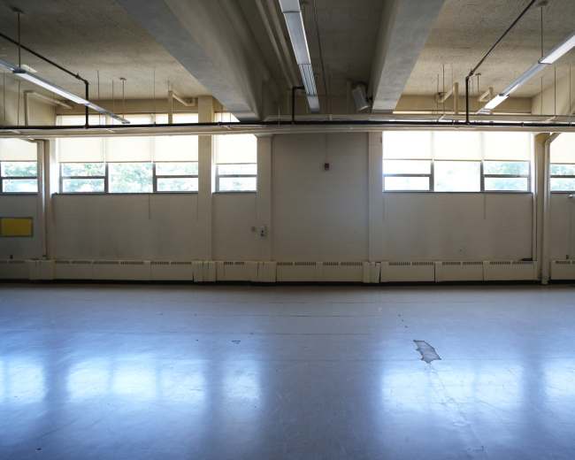 The image shows a spacious, empty room with large windows letting in natural light and a polished floor.
