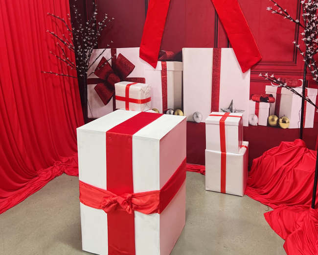 The scene features several large gift boxes wrapped in white paper with red ribbons, set against a backdrop of red drapery and decorations.