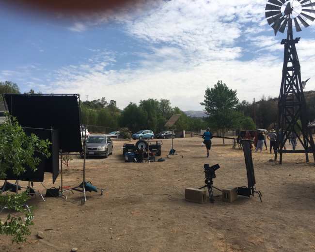 A film crew sets up equipment in a dusty outdoor location with a windmill in the background and vehicles parked nearby.
