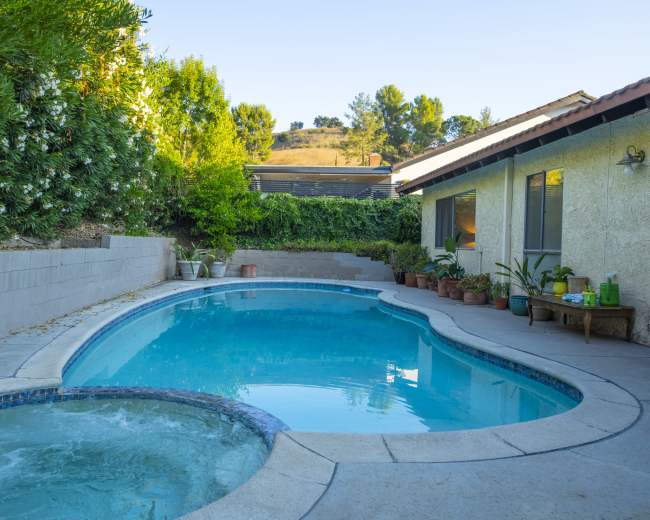 The image shows a backyard featuring a swimming pool and a hot tub, surrounded by lush greenery and various potted plants.