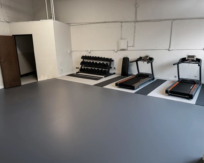 The image shows a gym interior with two treadmills, a set of dumbbells on a rack, and a door leading to another room.