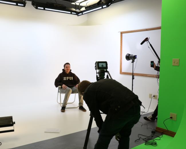 A seated individual in a hoodie is being filmed with a camera and lighting equipment in a studio environment.