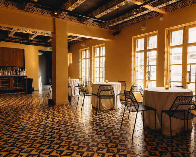 The image shows a restaurant interior with round tables covered in white tablecloths and black wire chairs arranged on a patterned tile floor.