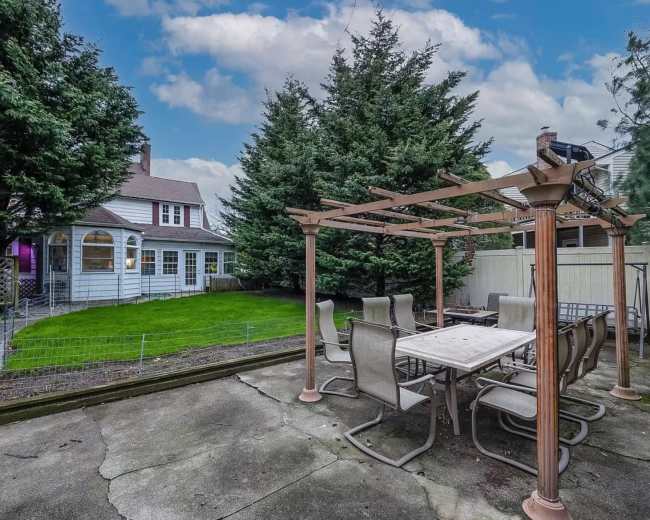 A stone patio with a pergola and outdoor dining set is situated in a backyard adjacent to a house and lined with tall trees.