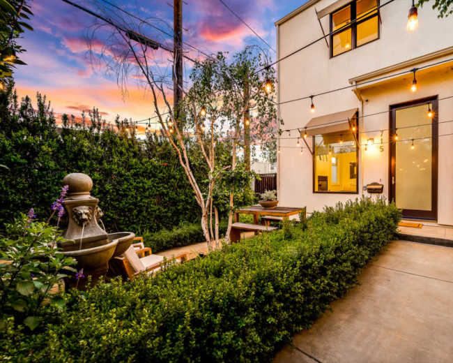 The image shows a backyard with a fountain, seating area, and string lights, set against a colorful sunset.