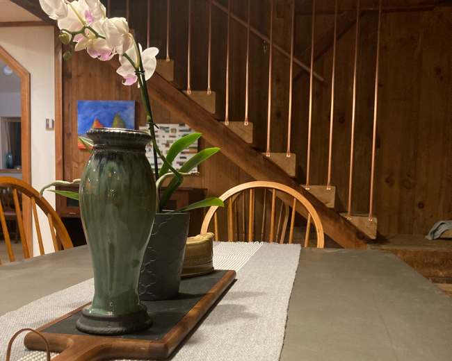 A dining table is set with a decorative vase containing an orchid, a wooden cutting board, and a textured runner in a rustic interior with a staircase in the background.