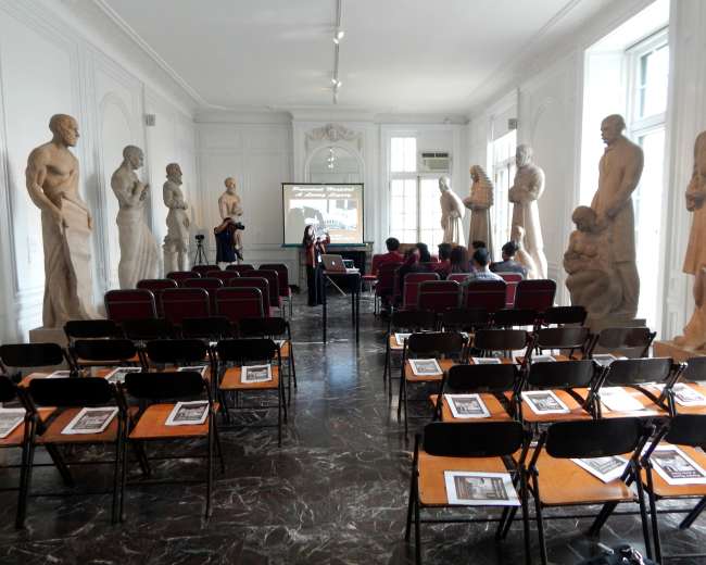 An art gallery with several large sculptures lining the walls and a row of empty chairs arranged in front of a screen.