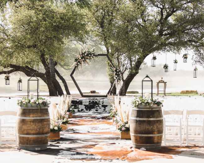The scene features a wedding ceremony setup with white chairs arranged in rows, a decorated hexagonal arch, and rustic barrels framing a carpet leading to the altar, all set under large trees.