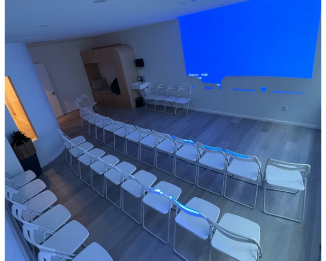 The image shows a minimalist seminar room with white chairs arranged in rows facing a blue projector screen.
