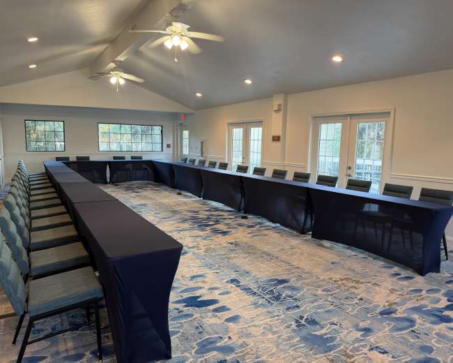 A large room features a U-shaped arrangement of tables covered with navy tablecloths, surrounded by chairs, in a well-lit space with large windows.