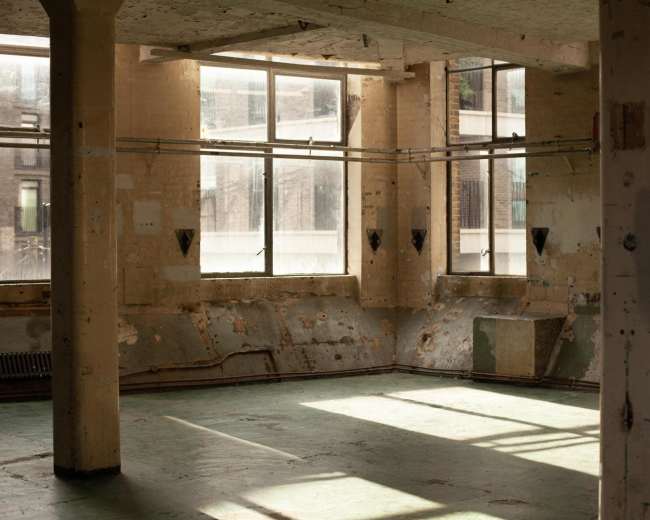 The image shows an empty, industrial room with large windows allowing light to enter, highlighting the worn walls and floor.