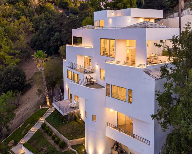 The image shows a modern white multi-level house with large windows, illuminated at dusk, situated on a hillside surrounded by greenery.