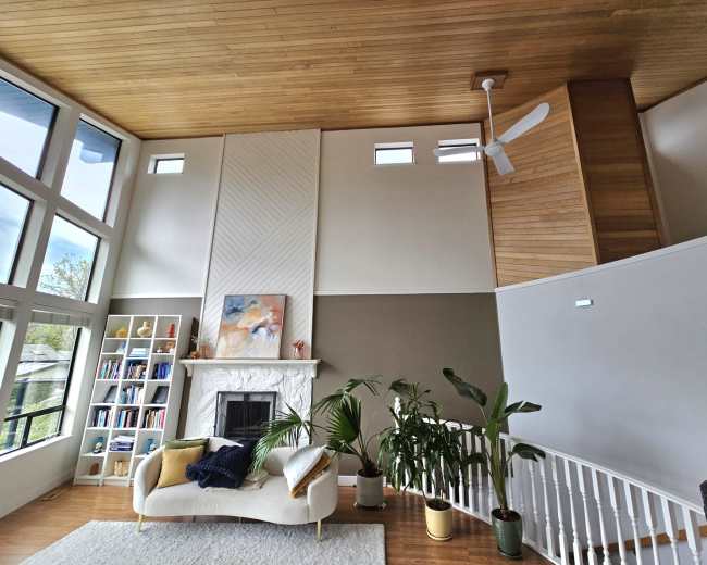 The image shows a modern living room with a wood-paneled ceiling, a light-colored couch, a fireplace, bookshelves, and several indoor plants.