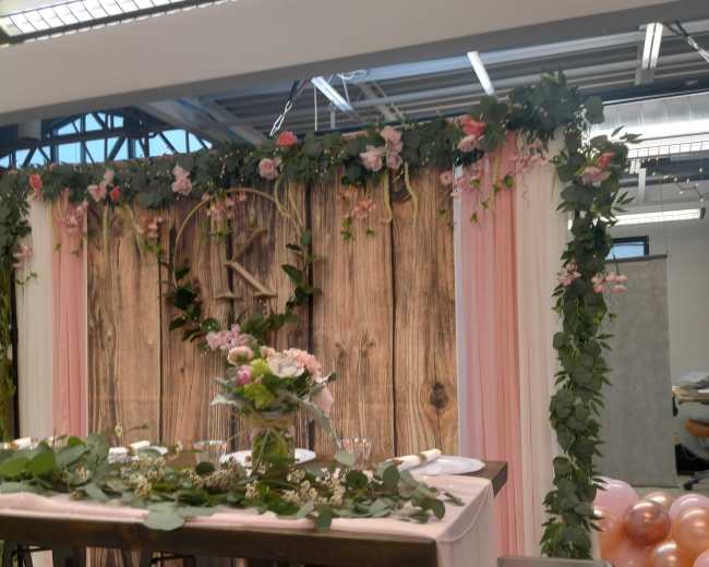 A decorated table set for an event, featuring a wooden backdrop adorned with flowers and greenery, surrounded by pastel balloons.