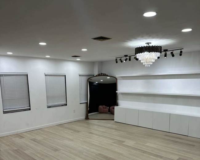 The image shows a spacious, well-lit room with wooden flooring, white walls, and shelves along one side, featuring a decorative chandelier and a mirror reflecting a darkened area.