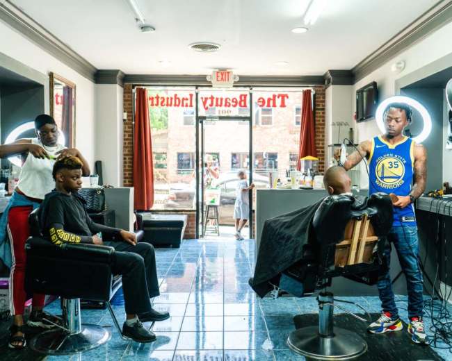 A barber styles a customer's hair while another individual sits in a chair, and a third person prepares for a haircut in a salon.