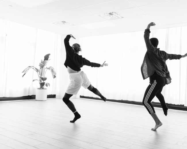 Two dancers perform in a brightly lit studio with large windows and a potted plant in the background.