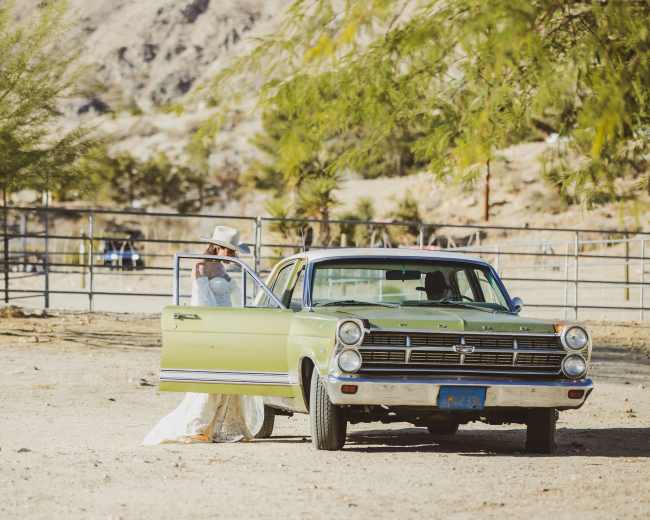 A woman in a white dress stands next to an open green sedan in a desert landscape.