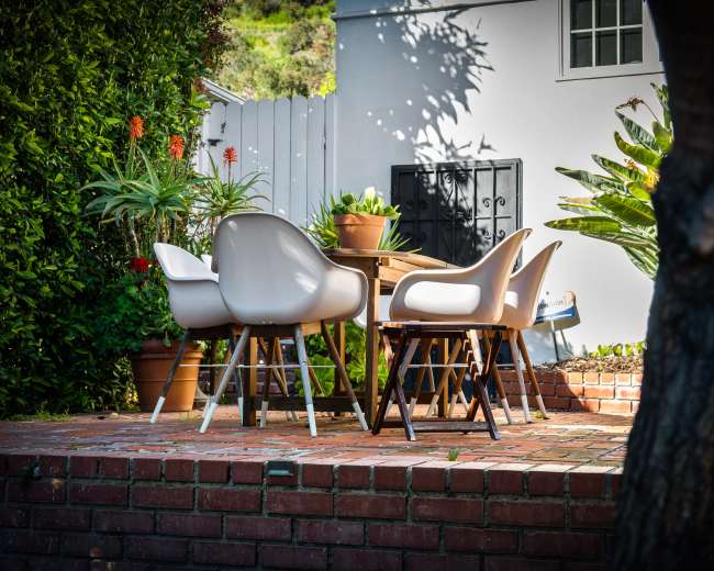 A set of modern chairs surrounds a wooden dining table on a brick patio, with potted plants and greenery in the background.