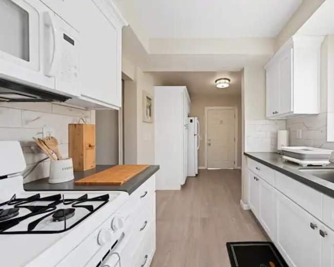The image shows a clean, modern kitchen with white cabinetry, a gas stove, and a hallway leading to a door at the end.