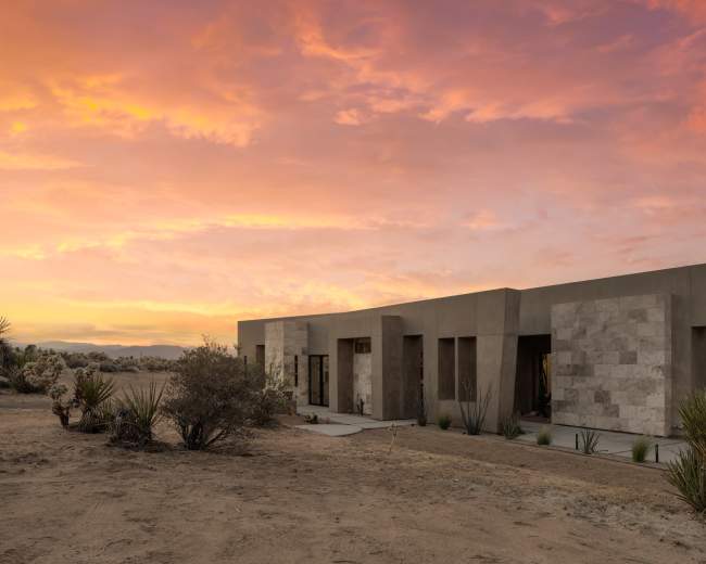 A modern building with a combination of concrete and stone finishes stands amid arid vegetation under a colorful sunset sky.