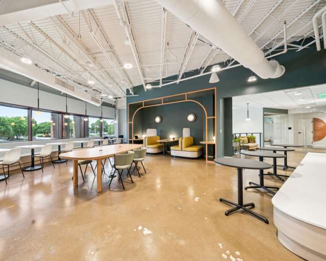 The image shows a modern office space with a variety of seating options, large windows, and a mix of tables and chairs arranged for collaborative work.