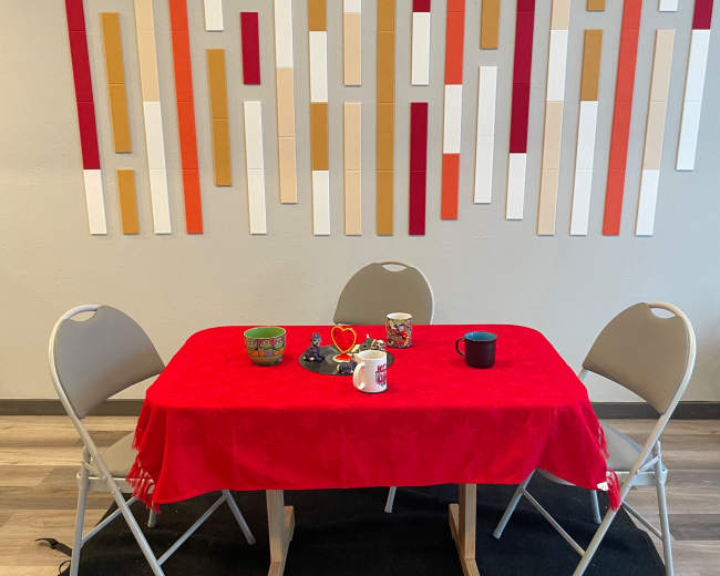A small table with a red tablecloth and three folding chairs is set against a wall decorated with vertical colored strips.